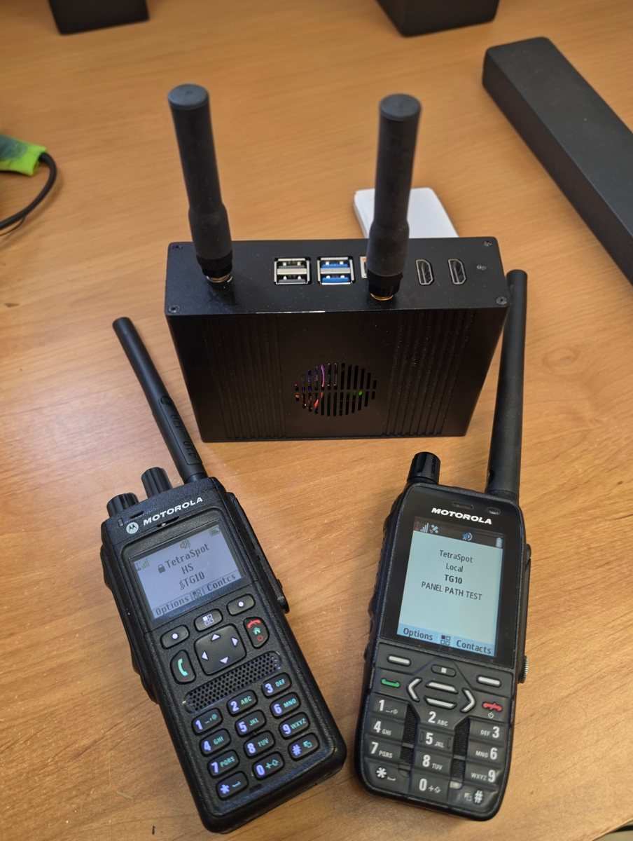 Stacja bazowa TetraSpot w czarnej obudowie z dwiema antenami, obok dwa radiotelefony Motorola zalogowane do grupy TG10